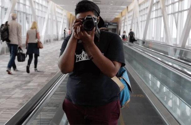 A person in a black t-shirt and maroon pants holds a camera. The person has dark brown skin and is looking into the camera to take a photo. They are standing on a mechanized walkway in an airport with various people walking back and behind the person.