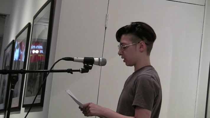 At a gallery, Sage Yockleson stands at a microphone reading from a piece of paper. They have light skin, short dark hair and glasses, and are wearing a plain t-shirt.