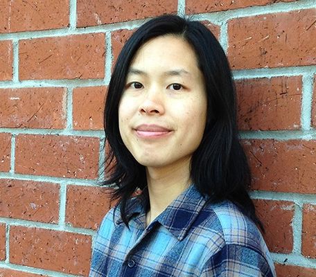 Bo Rittapa standing in front of a red brick wall. He has light skin, shoulder-length dark hair, and is wearing a blue plaid shirt. He has a calm and relaxed expression while slightly smiling at the camera.