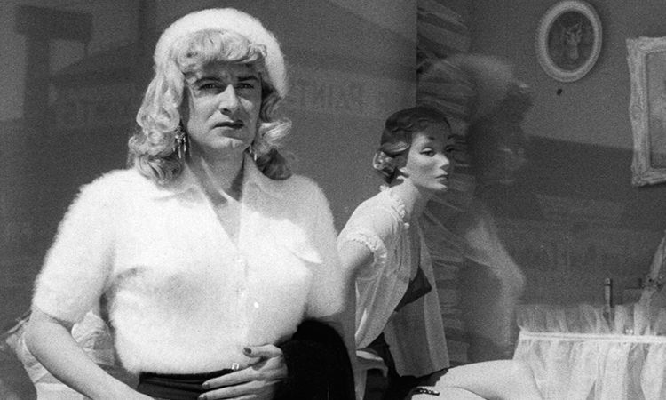 Black and white still from Glen or Glenda (1953) featuring the protagonist standing in front of a department store window.They have blonde curly shoulder length hair, light skin, dangly earrings, and a fuzzy white cap. They are wearing a fuzzy white blouse and black bottoms. Their right hand is resting on their stomach with dark nail polish on. 