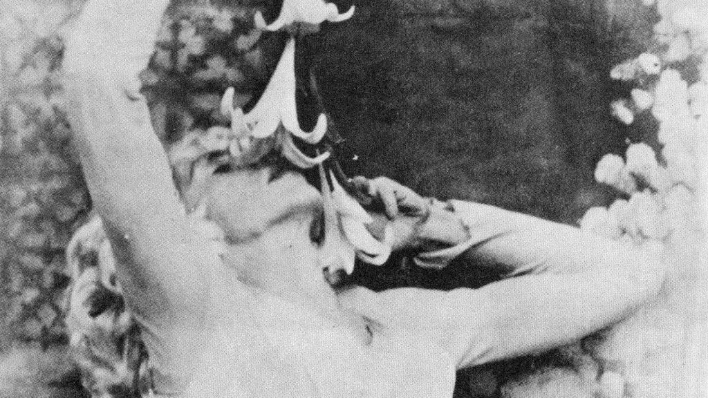 Black and white still from Flaming Creatures (1963). A character tilts their head back to smell a flower dangling over their face. They have light skin and blonde hair and a long sleeve white t-shirt. Their left hand is reaching up to hold the flower and the right is holding the flower to their face. The background is blurred out.