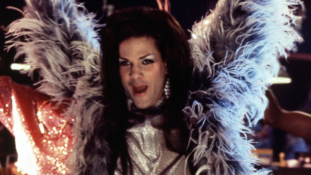 Still from Stonewall (1995). A character dressed in a feathered boa and shining dress raising their arms in the air. They have long straight brown hair and silver dangly earrings and light skin. 