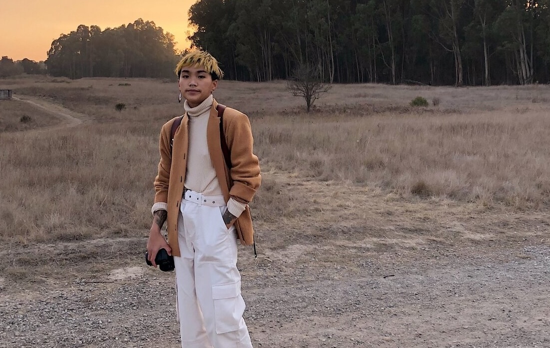 Sal Tran is nestled into a landscape with a road, a field of dried grass, trees, and a sunset sky. Sal Tran is a queer Vietnamese person with edgy short hair. They are holding a camera in their left hand and resting it against their left thigh. They are wearing a light brown blazer, light tan turtleneck, and white cargo pants. 