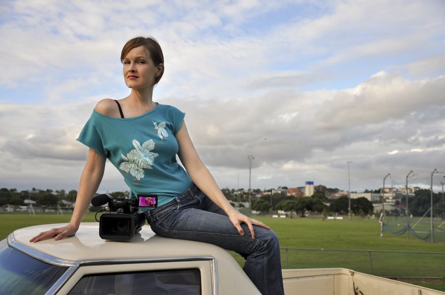 Still from Orchids: My Intersex Adventure (2011). Filmmaker Phoebe Hart sits on the roof of a pickup, camera by her side. They have light skin, wearing an off the shoulder short sleeve blue top with images of flowers on it in white.Their is black strap showing on their left shoulder and they are wearing denim jeans. They have short straight brown hair and posing with their right hand on their knee and the left hand rested on the hood of the car leaning towards the left hand that's propping them up. The background is a park with blue cloudy skies and distant trees.