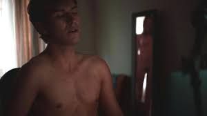 Still from @janabringlove (2016). Josef sits at his desk, unaware of the nude figure reflected in the mirror behind him. Josef is shirtless and the room is dark with the window curtains slightly open behind him letting in a small amount of light.